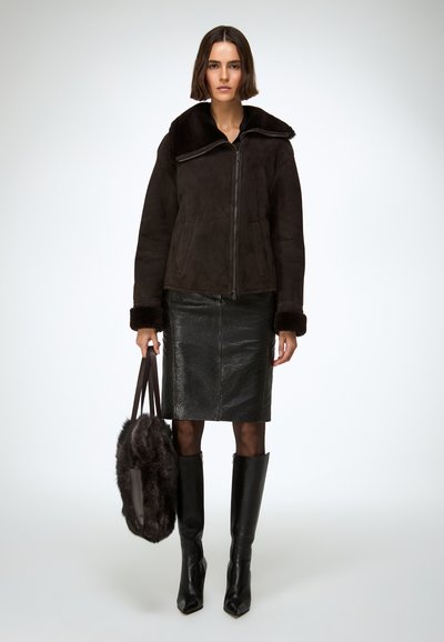 Brązowa kurtka z kożuchem o szerokim kołnierzu, zestawiona z czarną, teksturowaną skórzaną spódnicą oraz wysokimi, czarnymi botkami na obcasie sięgającymi do kolan. W dłoni futrzana torebka.