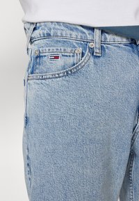Blugi din denim albastru deschis, cu o croială clasică, închidere cu nasture și un mic logo roșu, alb și albastru pe buzunarul din față.