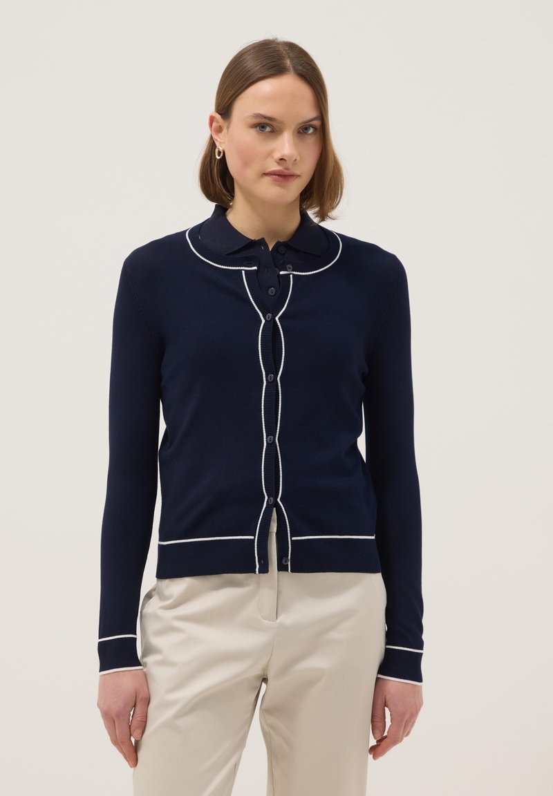 Frau trägt eine marineblaue Strickjacke mit weißen Besätzen und Knöpfen über einem marineblauen Poloshirt und beigen Hosen vor einfarbigem Hintergrund.