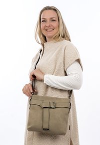 Femme souriante portant un gilet texturé beige et des manches longues blanches, tenant un sac à bandoulière taupe avec un détail en boucle.