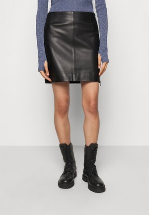 Persona vestida con una minifalda de cuero negra, botas de combate negras con cordones y una camiseta azul de manga larga con agujeros para el pulgar, de pie contra un fondo blanco.