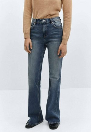 Person iført falmede blå bootcut-jeans, tan langærmet top og sorte sko, der står mod en ensfarvet lys baggrund.