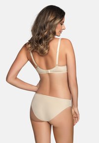 Mujer con cabello castaño ondulado vistiendo un sujetador beige y ropa interior a juego, de pie con una mano en la cadera, de espaldas sobre un fondo claro y liso.