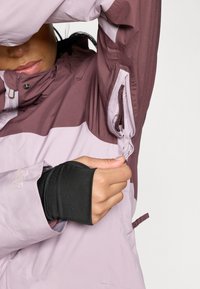 Vedenkestävä takki laventelin ja burgundin värissä, varustettuna vetoketjullisella taskulla, elastisilla hihansuilla ja korkealla kauluksella lisäsuojaksi.