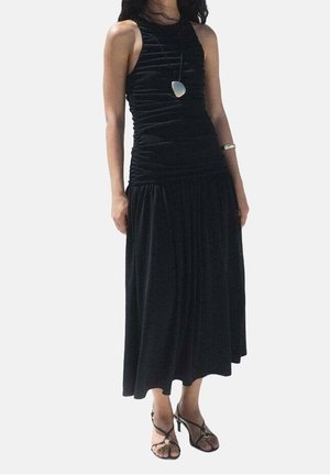 Femme portant une robe midi noire sans manches froncée, avec un collier pendentif argenté, un bracelet et des sandales à talons hauts noires à brides.