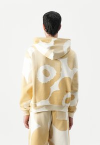 Gelb- und cremefarbener Hoodie mit großen Blumenmustern, aus weichem Baumwollmaterial und mit einer Kängurutasche. Verfügt über eine Kapuze mit Kordelzug und gerippte Bündchen.