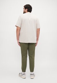 T-shirt beige à manches courtes associé à un pantalon de jogging vert olive et des baskets gris clair. Le tissu semble lisse, avec des poignets élastiques sur le pantalon.