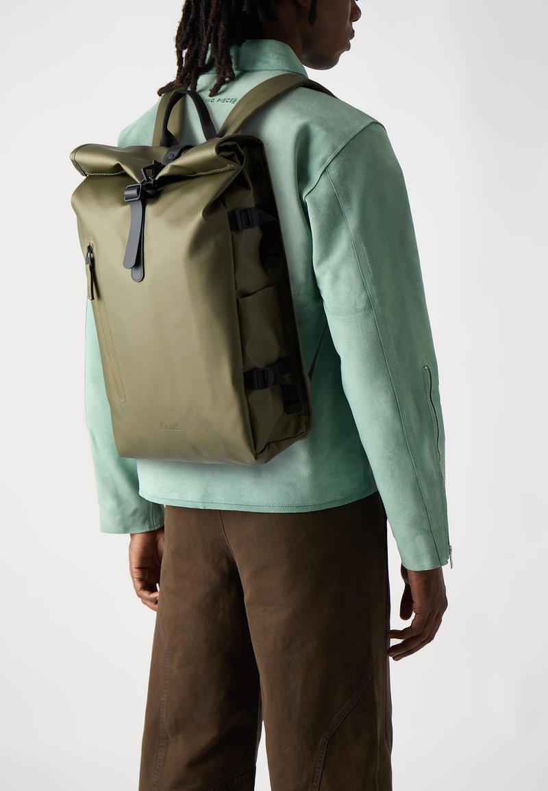 Sac à dos roulé de couleur vert olive, fabriqué en matériau imperméable, équipé de quincaillerie noire et de bretelles ajustables, porté par-dessus une veste verte menthe.