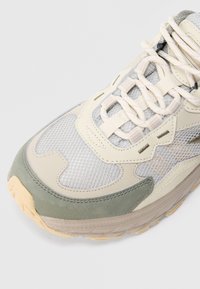 Sapato desportivo bege claro com painéis em rede, detalhes cinzentos e sola texturizada. Possui atacadores, colarinho almofadado e detalhes costurados.