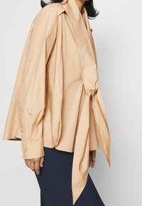 Femme portant une blouse beige ample avec des manches surdimensionnées et un long nœud devant, associée à une jupe bleu marine en côtes.