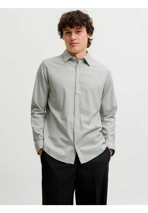 Jeune homme aux cheveux bouclés portant une chemise gris clair boutonnée et un pantalon noir, debout les mains dans les poches devant un fond uni.