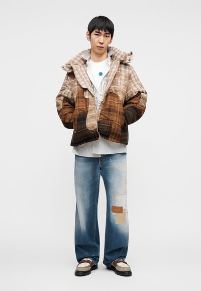 Πουκάμισο με κουκούλα σε καρό μοτίβο σε καφέ και μπεζ αποχρώσεις, συνδυασμένο με ανοιχτόχρωμο τζιν παντελόνι με επιδιορθώσεις και λευκό T-shirt με γραφικό σχέδιο.