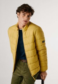 Chaqueta acolchada amarilla con diseño de acolchado, cuello alzado y cierre de cremallera, llevada sobre un suéter azul oscuro y pantalones verde oliva.