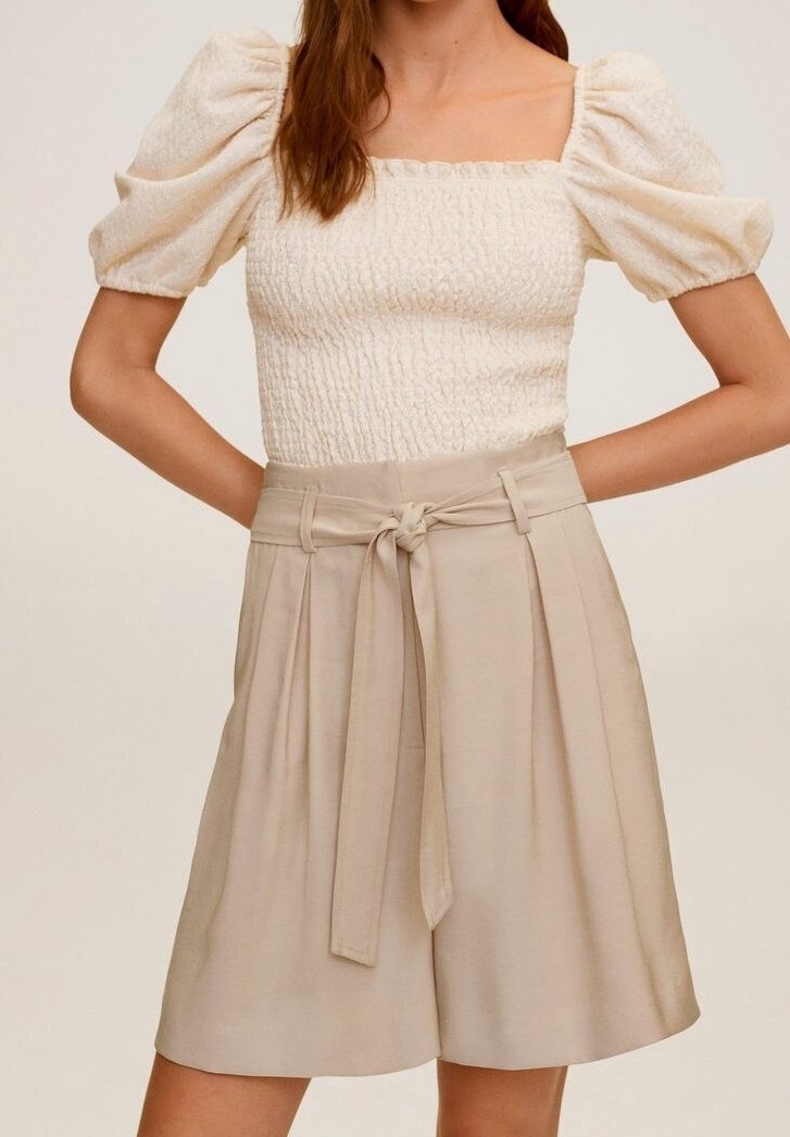 Femme portant un haut blanc texturé à manches bouffantes, associé à un short beige taille haute noué à la taille avec une ceinture en tissu.