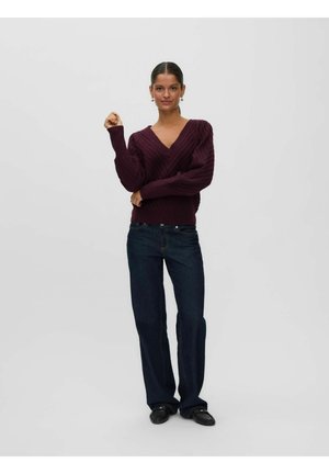 Kvinde iført en mørk lilla V-hals sweater, mørkeblå jeans og sorte loafers, stående med den ene arm hævet og den anden foldet over taljen.