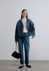 Μπουφάν από τζιν με διακοσμητικές φούντες, φορεμένο πάνω από λευκό πουλόβερ. Συνδυασμένο με μπλε τζιν, μαύρες μπαλαρίνες και πλισέ μαύρη τσάντα χειρός.