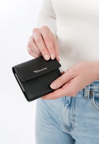 Manos sosteniendo y abriendo una pequeña cartera negra de cuero con un logo plateado de "Tamaris", persona vestida con jeans claros y una blusa de manga larga blanca.