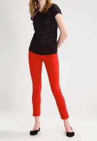 Černý top s krátkým rukávem a mřížkovaným vzorem, který je kombinován s těsnými červenými kalhotami a černými balerínami, č ímž vytváří kontrast barev a textur.