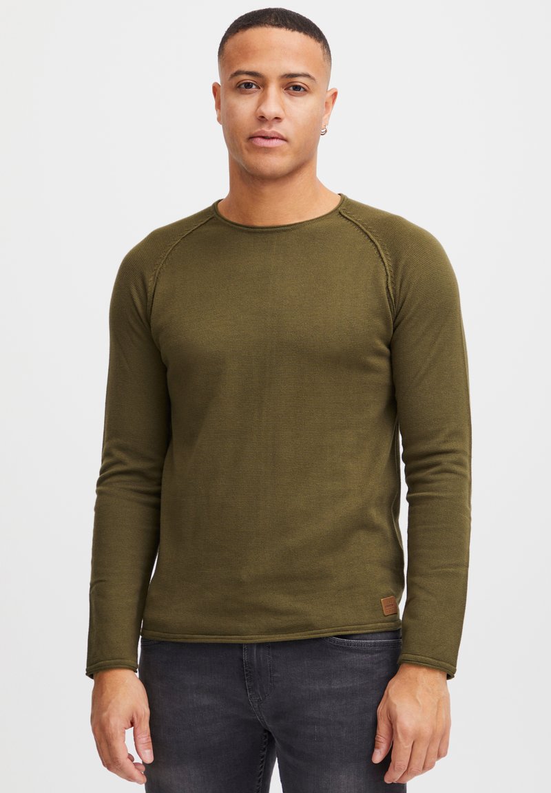 Chemise à manches longues vert foncé avec une finition texturée, col rond et manches raglan. Petit patch de marque près de l'ourlet.