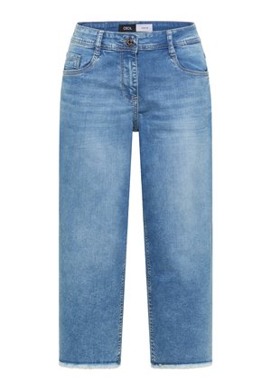 Frau mit gelber Kappe, schwarzem kurzärmligem Hemd mit Knöpfen, kurzen blauen Jeans, beigen Turnschuhen und trägt eine gesteppte hellgrüne Tragetasche.