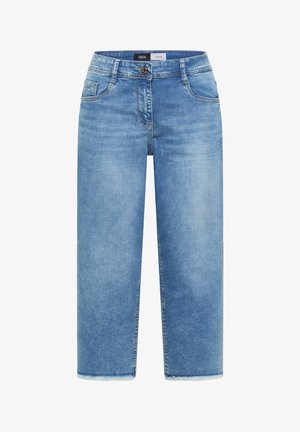 Kvinde iført gul kasket, sort kortærmet skjorte med knapper, korte blå jeans, beige sneakers og bærer en quiltet lysegrøn skuldertaske.
