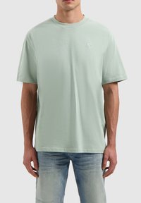 Lichtgroene katoenen t-shirt met een ronde hals, korte mouwen en een klein geborduurd logo op de borst, gecombineerd met lichtblauwe jeans.