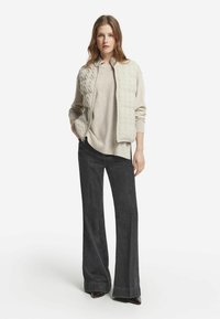 Junge Frau in einer beigen Strickjacke mit Reißverschluss, leichtem Pullover, weiten schwarzen Jeans und dunklen spitzen Schuhen, stehend vor weißem Hintergrund.