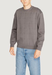 Maglione grigio a maglia con un motivo a griglia testurizzato, collo rotondo e polsini a coste. Abbinato a jeans di denim azzurri chiari.