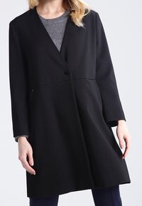 Černý dlouhý kabát s texturovaným povrchem, výstřihem do V, zapínáním na jeden knoflík a bočními kapsami. Má dlouhé rukávy a volný střih.