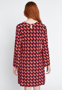 Langsleeve jurk met een kleurrijk geometrisch patroon in rood, roze en zwarte tinten. De keyhole achterkant voegt visuele belangstelling toe.