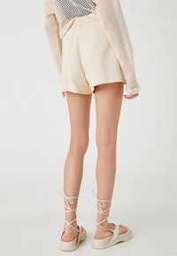 Pantalones cortos de algodón beige con cintura elástica, combinados con sandalias claras con tiras cruzadas. El fondo es blanco y neutro.