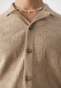 Maglione beige a collo, con un motivo testurizzato di forme circolari e tre bottoni sul davanti.