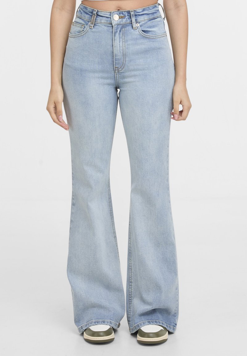 Femme portant un jean évasé taille haute bleu clair associé à des baskets blanches et vert olive, debout devant un fond uni.