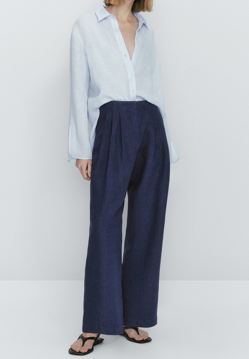 Personne portant une chemise bleu clair boutonnée, rentrée dans un pantalon taille haute à jambes larges bleu foncé, avec des sandales noires.