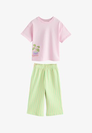 Roza majica kratkih rukava s grafikom nasmijanog biljnog lonca u kombinaciji sa širokim hlačama s okomitim prugama u zelenoj i breskvastoj boji.