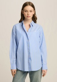 Jasnoniebieska koszula w paski w kolorze białym, zapinana na guziki, z długimi rękawami, klasycznym kołnierzykiem i małym haftowanym logo na klatce piersiowej.