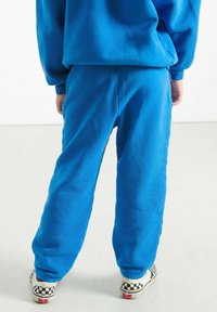 Cilvēks ar ziliem sporta biksēm un atbilstošu džemperi, stāv ar rokām blakus, uzvilcis melnbaltas šaha laukuma raksta slip-on kurpes.