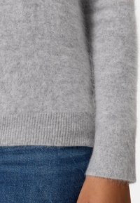 Maglione grigio peloso con orlo e polsini a coste, realizzato in un materiale morbido; indossato sopra un jeans blu, con una texture liscia e pelosa.