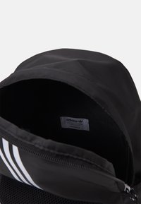 Černý batoh s hladkým povrchem, s třemi bílými pruhy, se zipovou přihrádkou a štítkem s nápisem "adidas PRIMEGREEN."