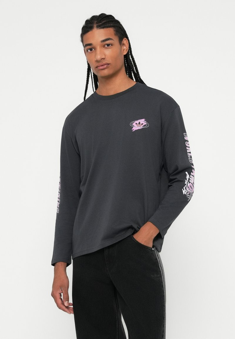 Mann mit langen geflochtenen Haaren, der ein dunkles Langarmshirt mit pinkem Logo auf der Brust und Text auf den Ärmeln trägt, mit der Hand in der Tasche seiner schwarzen Hose.