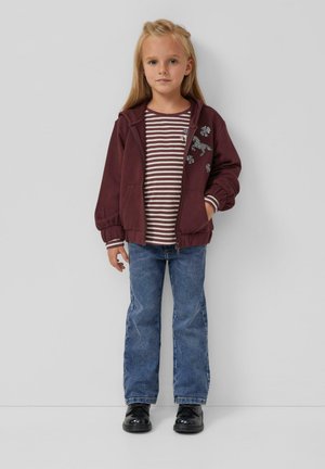 Bordeauxfarbener Reißverschluss-Hoodie mit floralen Verzierungen, gestreiftem Langarmshirt, blauen Jeans und schwarzen Lackschuhen. Gerader Schnitt und gekrempelte Ärmel.
