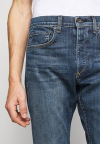 Gros plan d'une personne portant un jean en denim bleu et une chemise blanche, montrant la taille, les poches et la main gauche détendue sur le côté.