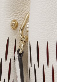 Detail bílé kožené tašky s zlatými prvky, klipem a červenými výřezy po straně. Texturovaný povrch nabízí hmatovou kvalitu.