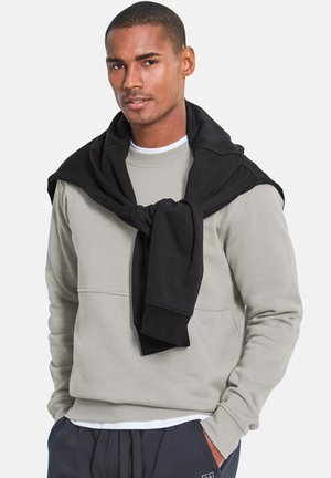 Junger Mann trägt beige Sweatshirt mit einem schwarzen Sweatshirt um die Schultern gebunden und navyblaue Hose, steht vor einem einfarbigen Hintergrund.