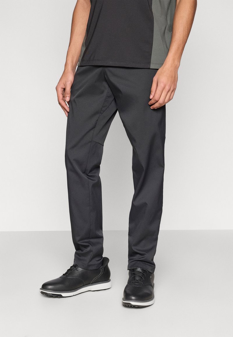 Ανδρικό μαύρο παντελόνι από ανθεκτικό, λείο υλικό, με στενή εφαρμογή και διακριτικές ραφές, συνδυασμένο με μαύρα επίσημα παπούτσια.