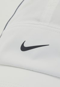 Hvid kasket lavet af letvægtsstof, med et sort Nike swoosh-logo på forsiden. Glat tekstur med syede sømme.