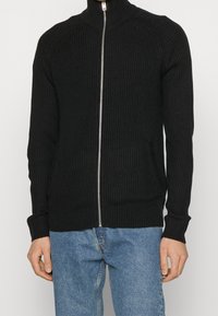 Svart ribbad stickad zip-up-tröja med hög krage, sidofickor och långa ärmar. Texturen är mjuk och har en figurnära design.
