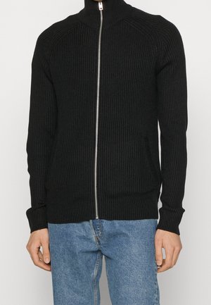 Maglione nero a coste con zip e collo alto, tasche laterali e maniche lunghe. La texture è morbida, con un design aderente.