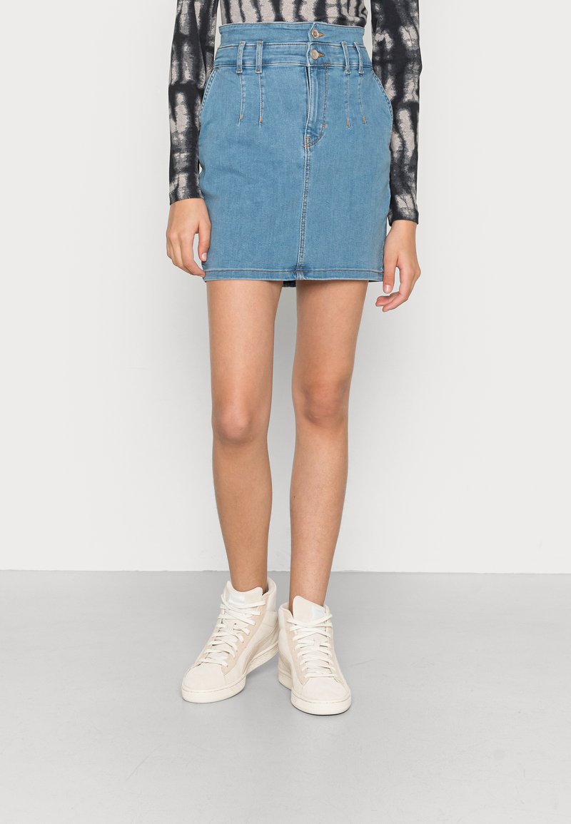 Saia mini de ganga em azul claro com cintura alta, apresentando dois botões frontais, bolsos laterais e uma textura suave. Combinada com ténis brancos de cano alto.