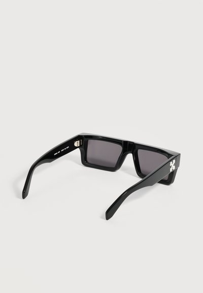 Lunettes de soleil rectangulaires noires avec des montures épaisses et des verres foncés, montrant un petit logo blanc sur la branche, présentées sur fond blanc.